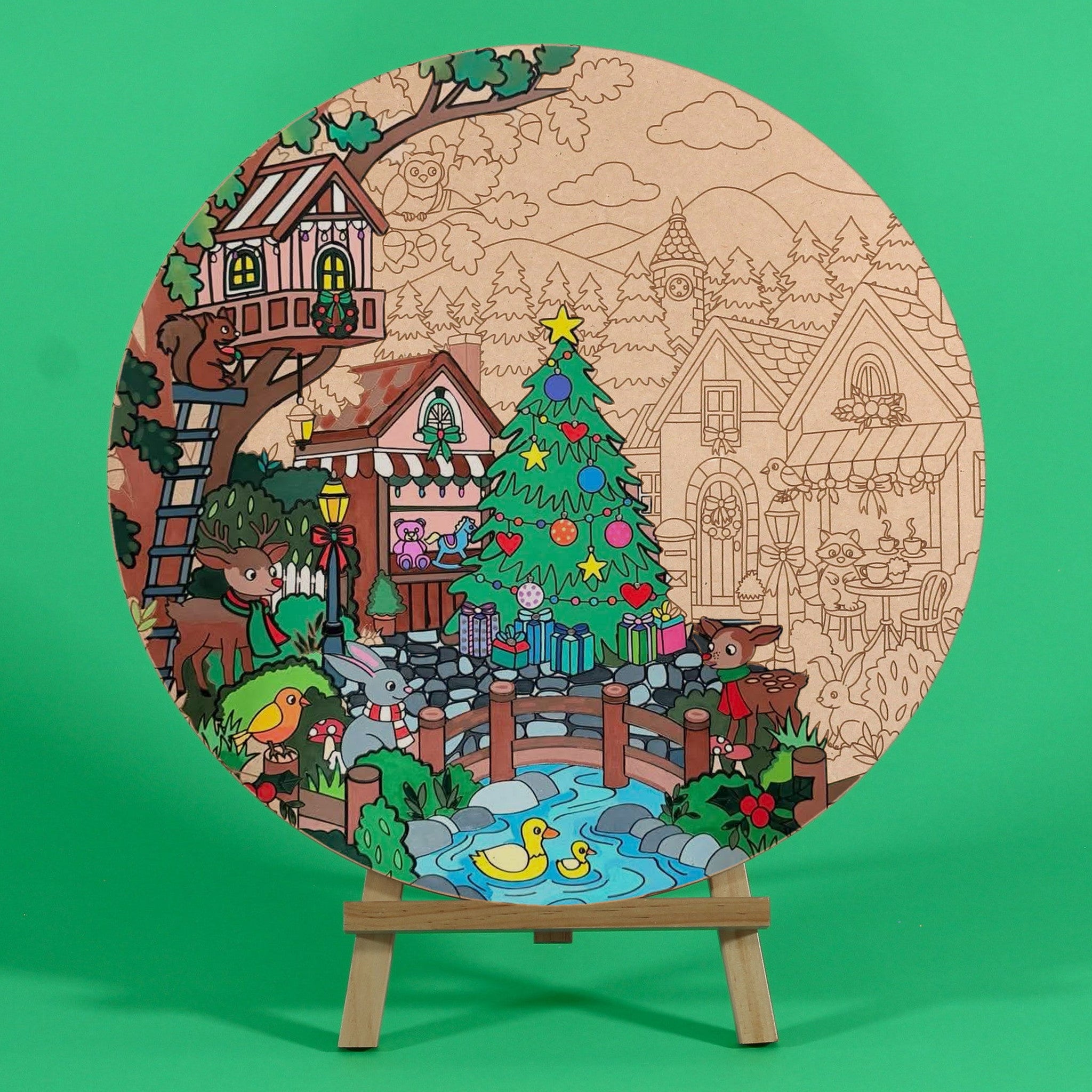 A Woodland Christmas 400mm engraved MDF art board by Hueray, half coloured by a customer, showing a festive woodland village with a treehouse, Christmas tree and stream, on a wooden easel on a green background