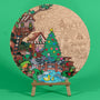 A Woodland Christmas 400mm engraved MDF art board by Hueray, half coloured by a customer, showing a festive woodland village with a treehouse, Christmas tree and stream, on a wooden easel on a green background