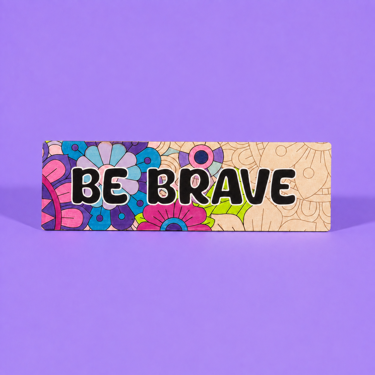 A rectangular decorative sign with the words 'BE BRAVE' in bold black letters. The left side features a colorful retro floral pattern in pink, blue, purple, and green, while the right side has a beige background with light brown line-art doodles. Showing partly coloured in 