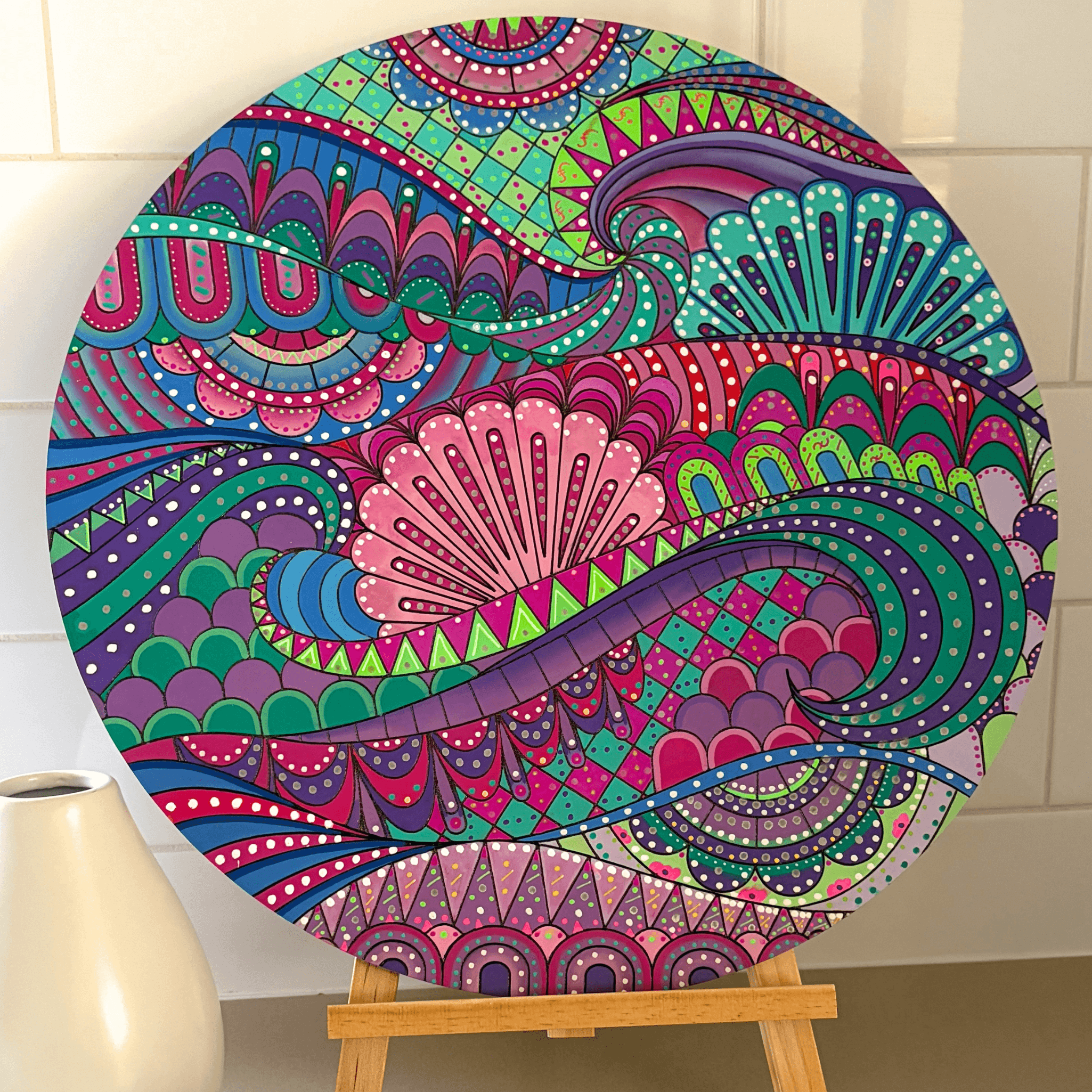 A round canvas print featuring a vibrant, intricate mandala and paisley pattern in shades of pink, purple, teal, green, and blue, displayed on a wooden easel against a white tiled wall.