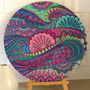 A round canvas print featuring a vibrant, intricate mandala and paisley pattern in shades of pink, purple, teal, green, and blue, displayed on a wooden easel against a white tiled wall.