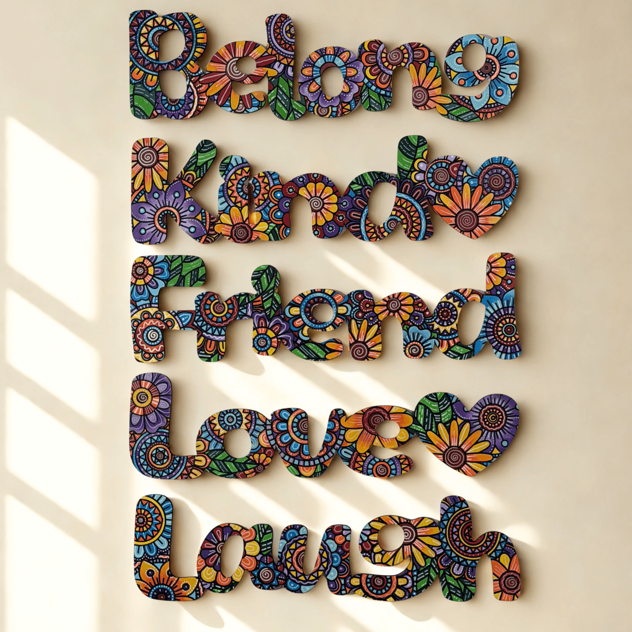 Multicolor decorative wall art spelling out 'Belong Kind Friend Love Laugh' in engraved floral-patterned letters, mounted on a beige wall with soft window-shadow lighting.