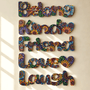 Multicolor decorative wall art spelling out 'Belong Kind Friend Love Laugh' in engraved floral-patterned letters, mounted on a beige wall with soft window-shadow lighting.