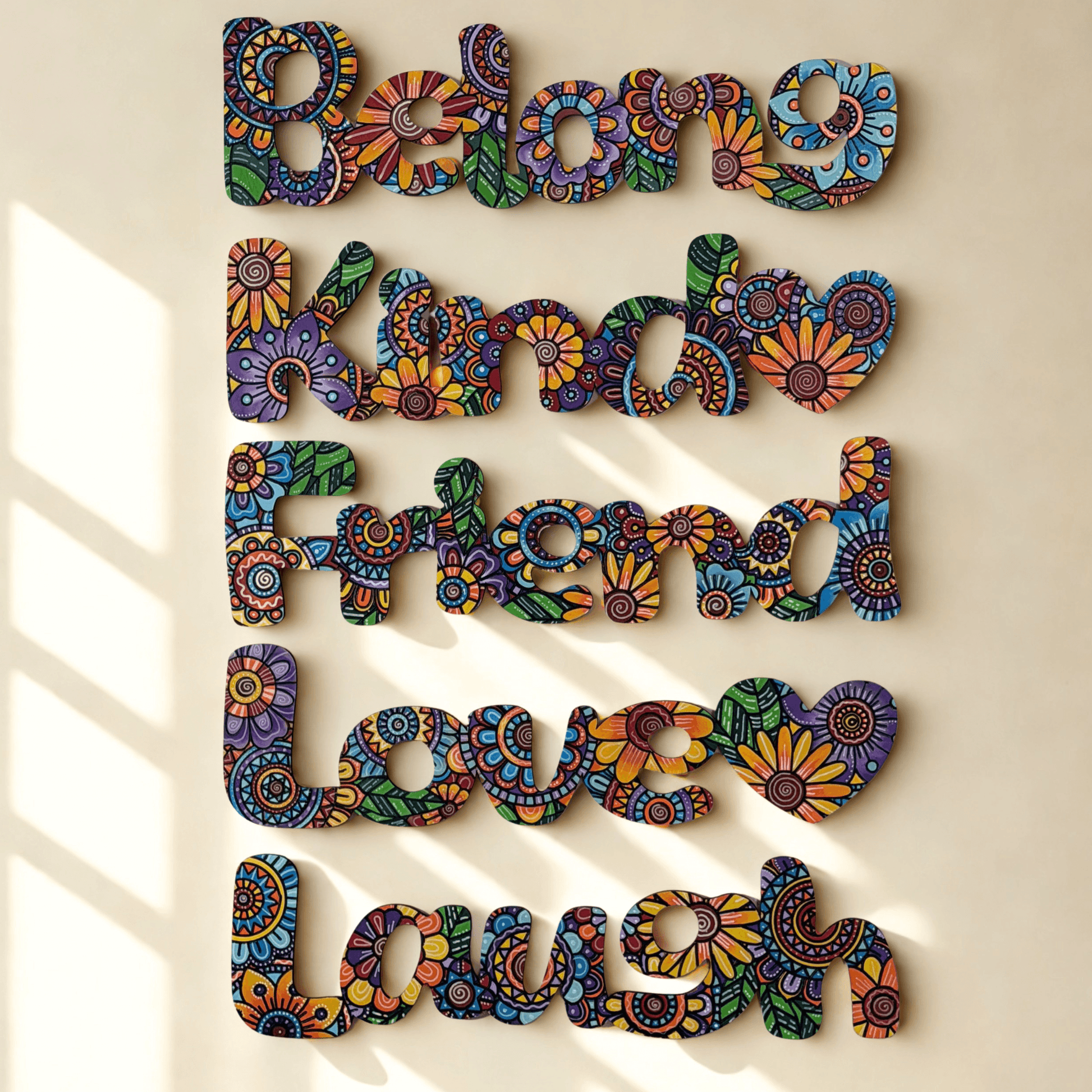 Multicolor decorative wall art spelling out 'Belong Kind Friend Love Laugh' in engraved floral-patterned letters, mounted on a beige wall with soft window-shadow lighting.