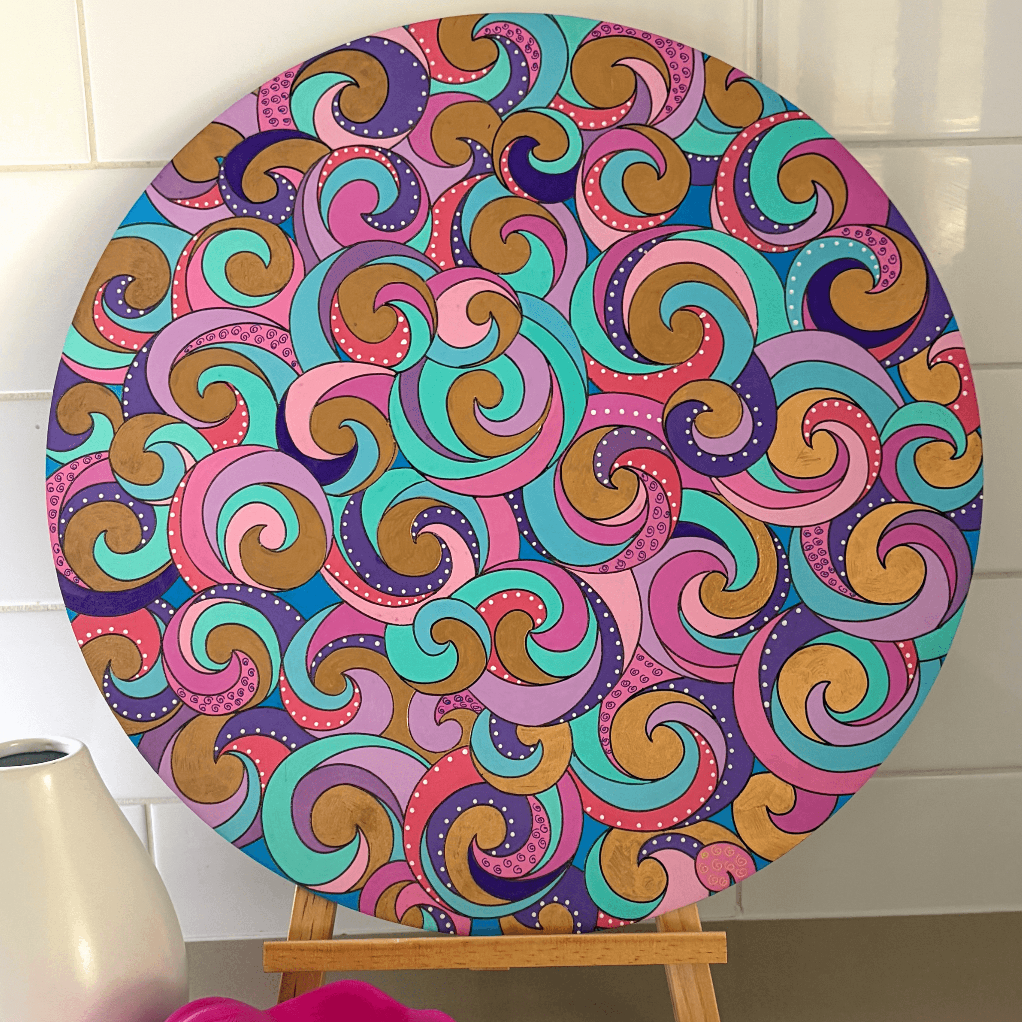 A round canvas print featuring a colorful retro swirl pattern, displayed on a wooden easel against a white tiled wall.