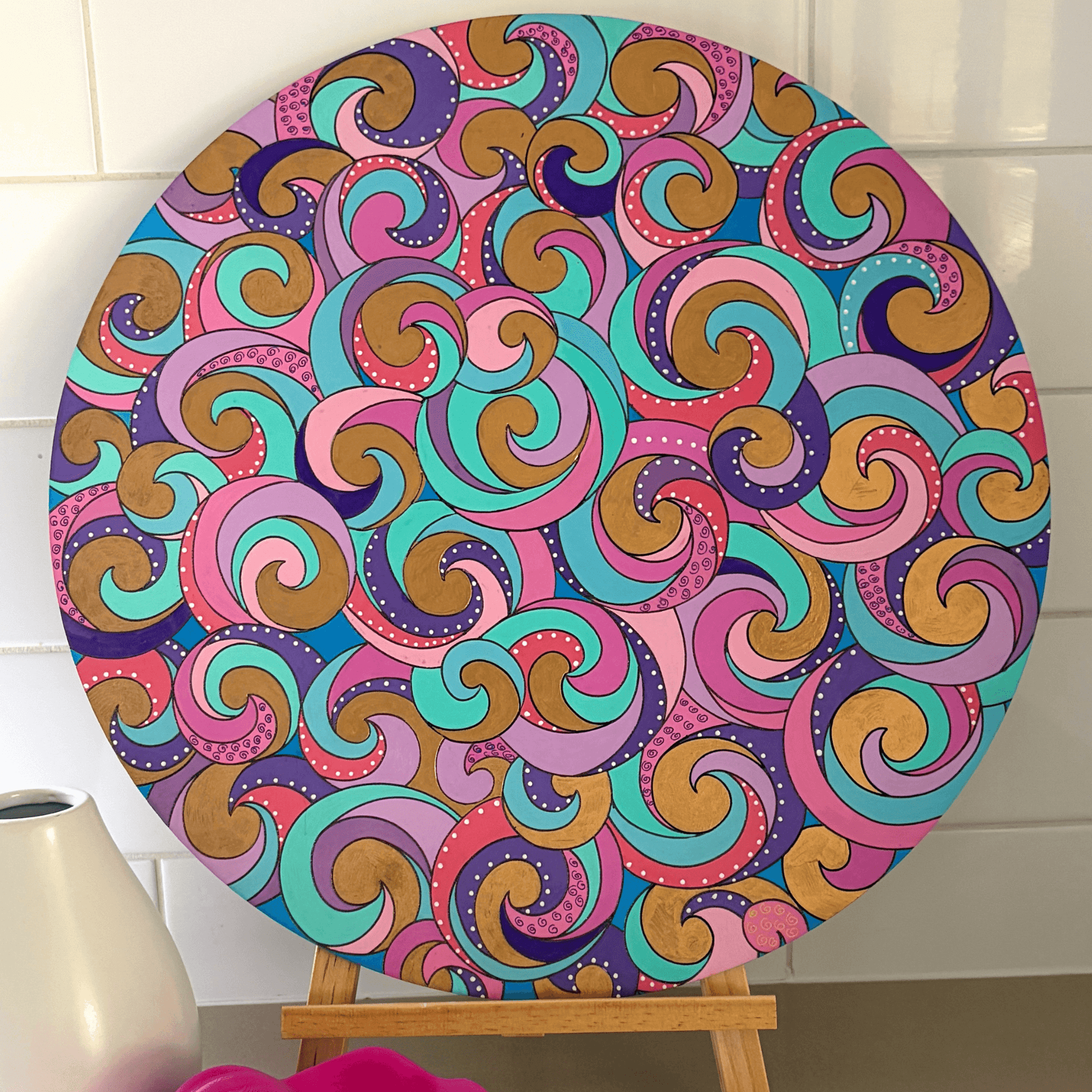 A round canvas print featuring a colorful retro swirl pattern, displayed on a wooden easel against a white tiled wall.