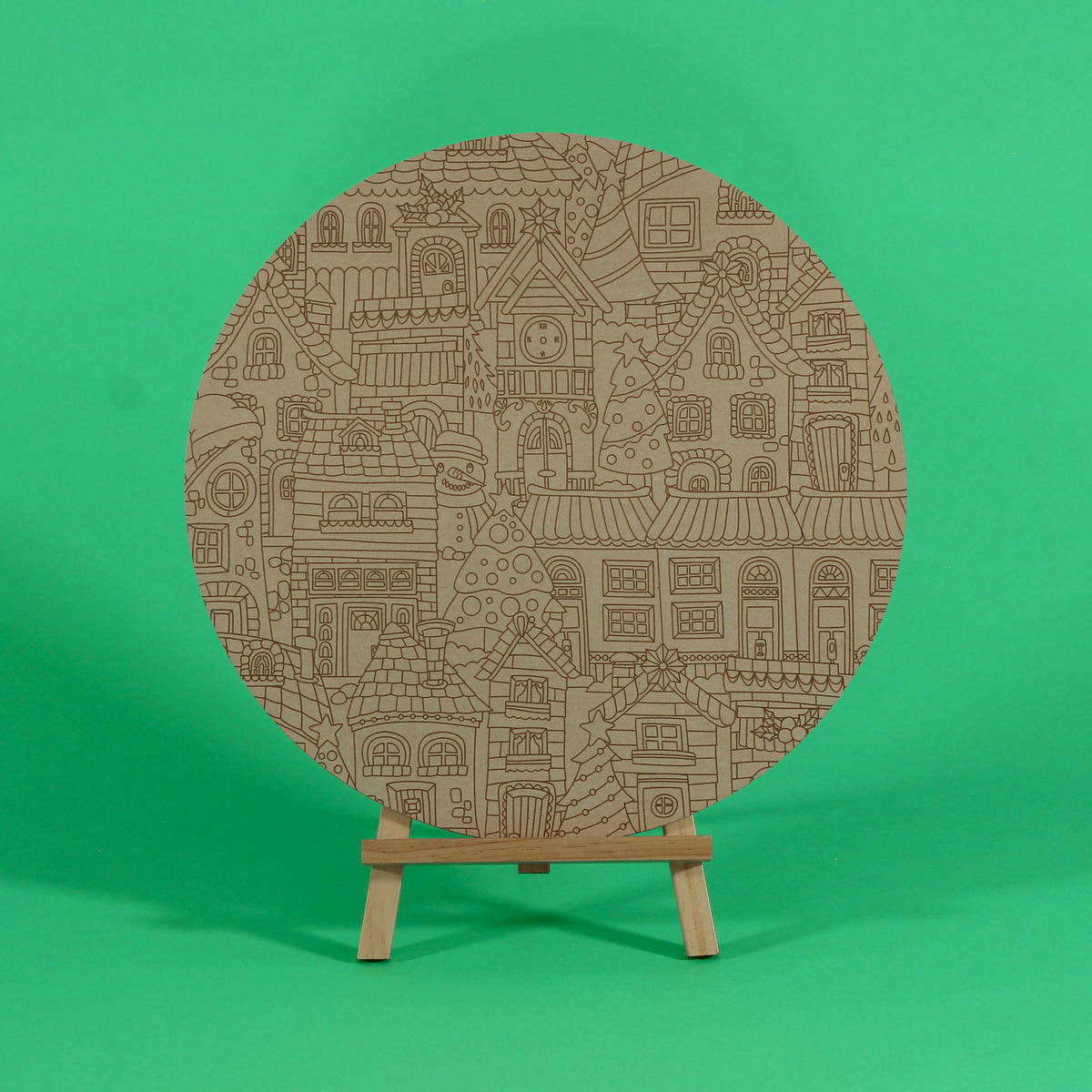Uncoloured Twinkle Town 400mm round MDF board on an easel, engraved with a Christmas village of houses, a clock tower and Christmas trees