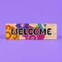 A rectangular decorative sign with the word “WELCOME” in large black letters over a colorful floral and geometric pattern, photographed against a solid purple background.