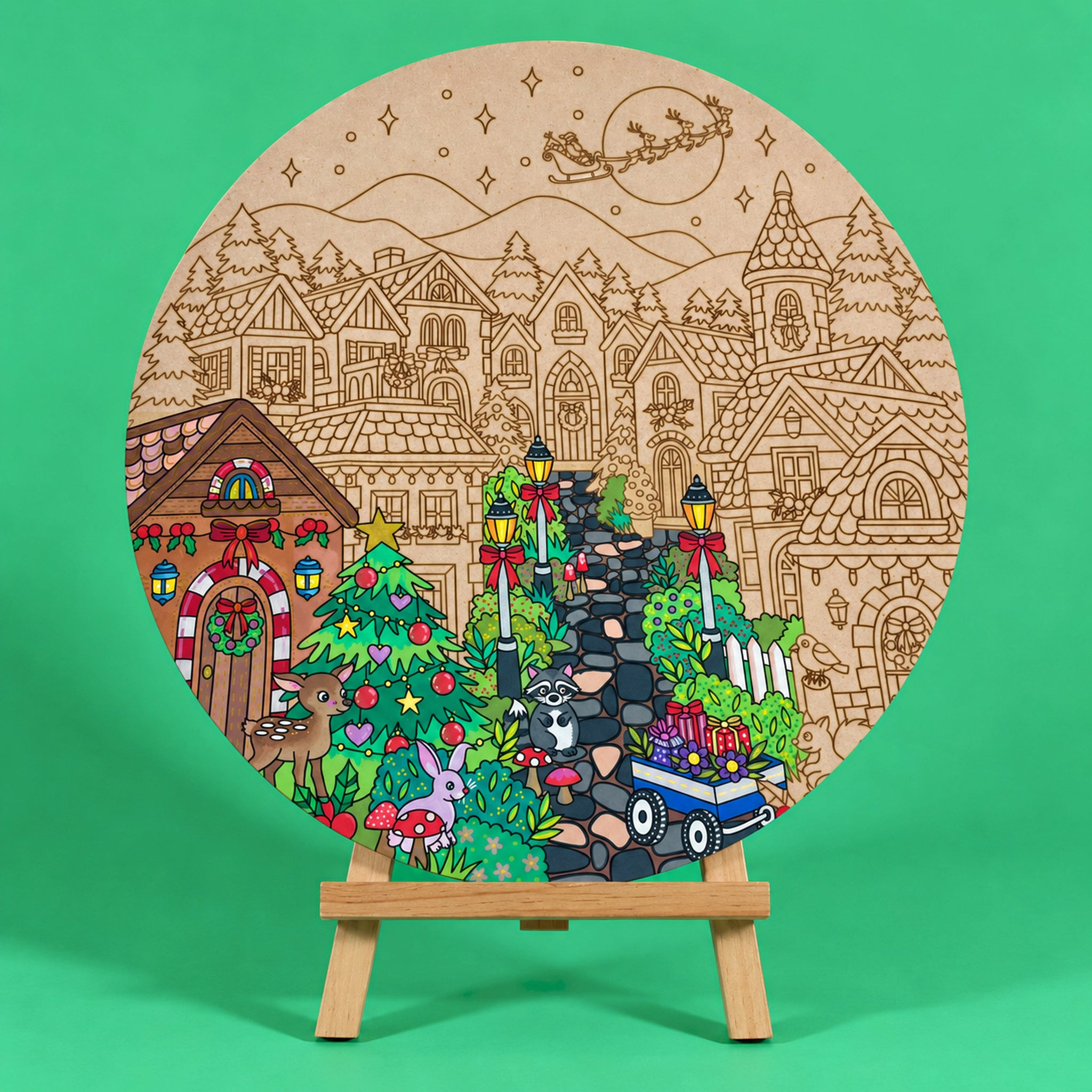 Mistletoe Lane 400mm round Christmas art board on an easel, partly coloured, with storybook houses, a Christmas tree, a deer, a rabbit and Santa's sleigh over the moon