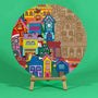 Twinkle Town 400mm round Christmas art board on an easel, half coloured in rainbow shades, showing village houses, a clock tower and Christmas trees